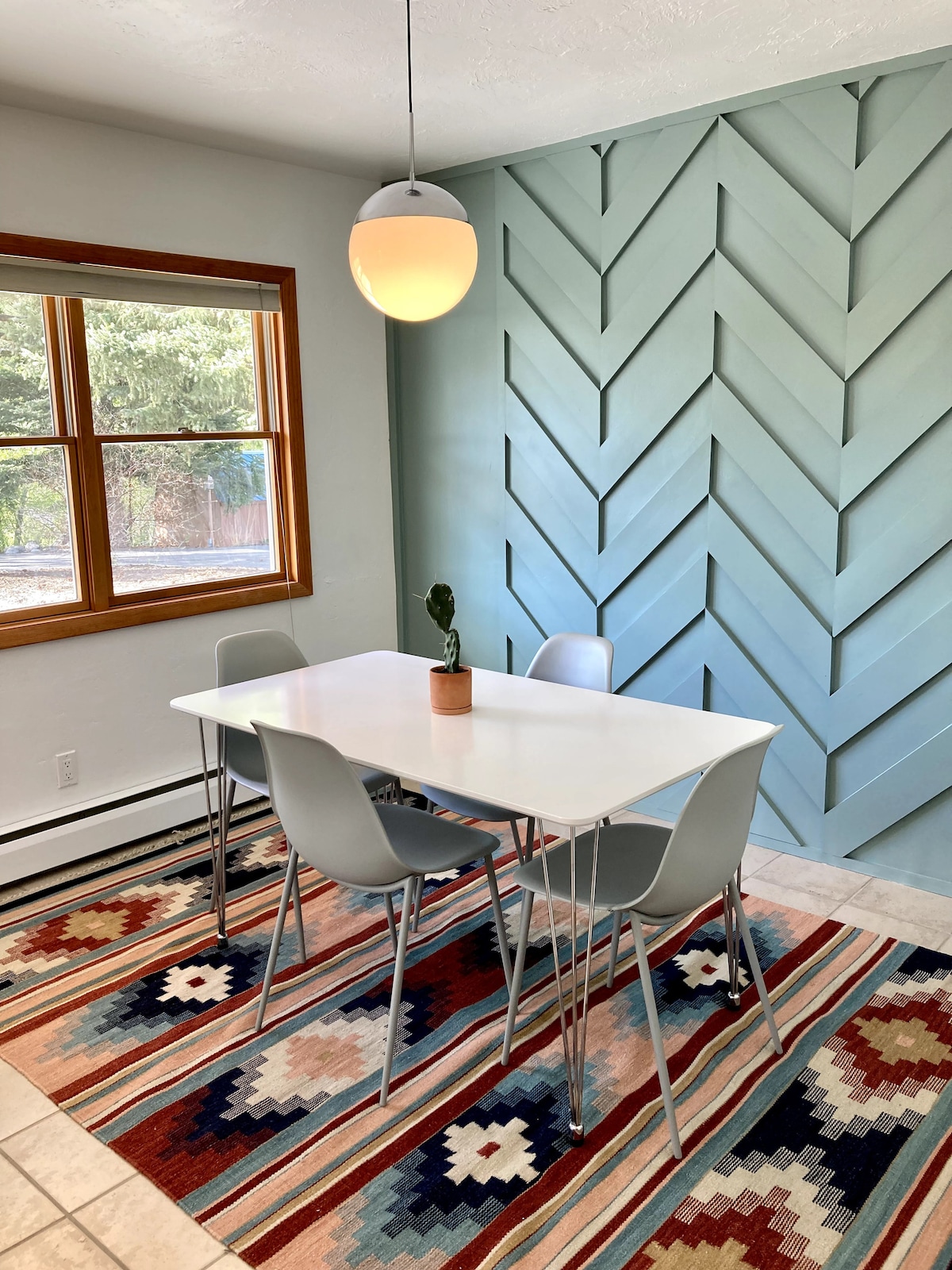 A dining area features a modern white table surrounded by four sleek chairs. A colorful patterned rug adds warmth to the space, while a pendant light illuminates the area. A decorative plant sits on the table, and a large window provides natural light.