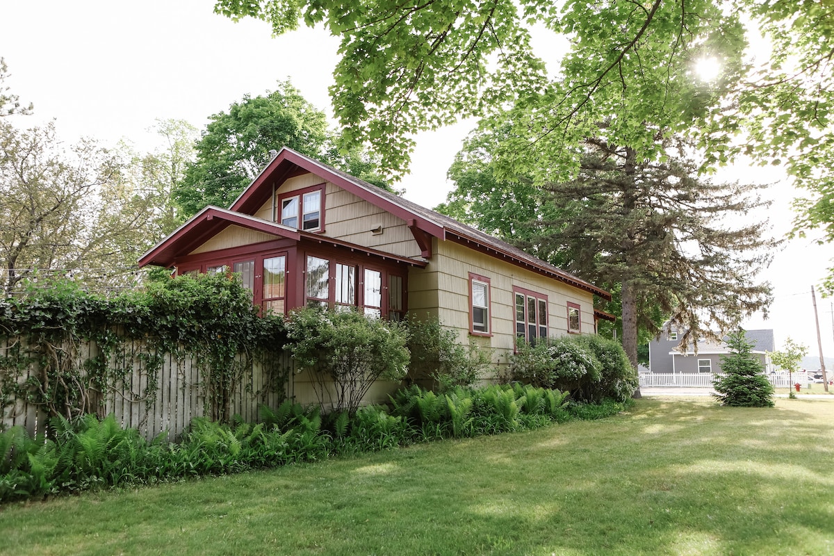 A charming craftsman home is nestled among lush greenery, featuring a gabled roof with red trim. Large windows invite natural light, while the surrounding gardens are filled with vibrant ferns and well-kept hedges, enhancing the serene outdoor ambiance.