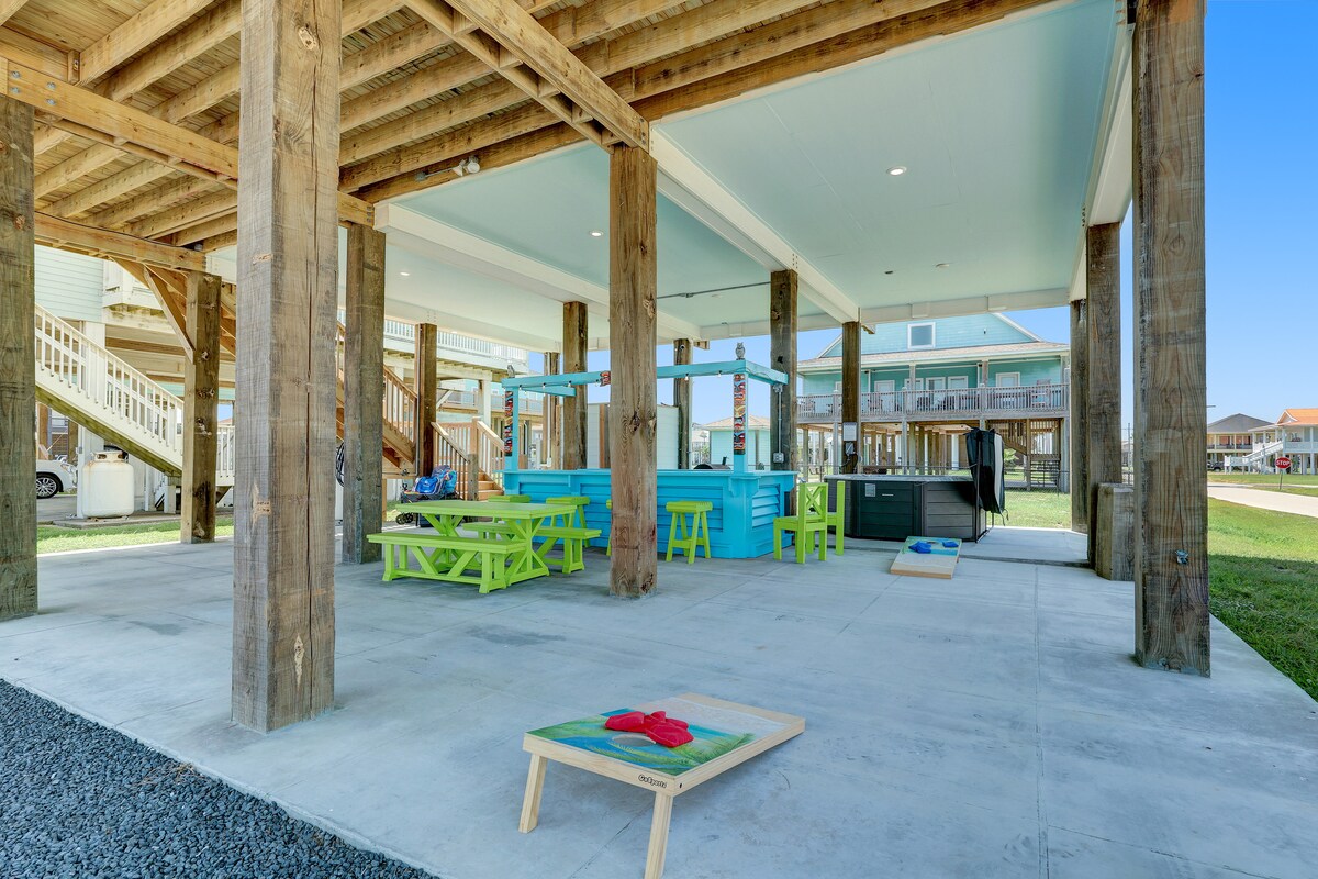 An open, shaded space is illuminated by natural light, featuring a picnic table set in vibrant green and additional seating in blue. A hot tub is located discreetly in the corner, while sand games are visible on the ground, providing options for outdoor entertainment.