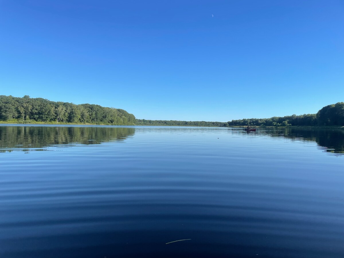 A calm river is surrounded by lush green trees on both banks, creating a serene natural setting. The water’s surface reflects the clear blue sky, enhancing the tranquil atmosphere. The gentle ripples across the water suggest a peaceful environment, ideal for relaxation.