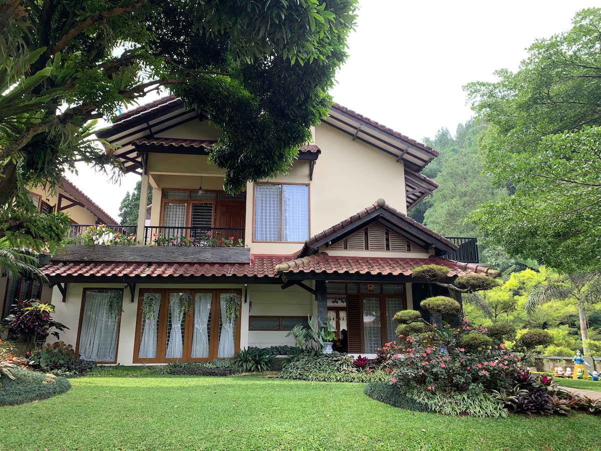 The villa features a two-story structure with a traditional roof and multiple large windows. Lush green lawns are surrounded by colorful flower beds and meticulously shaped shrubs, creating a serene outdoor space. Towering trees provide additional shade and natural beauty.