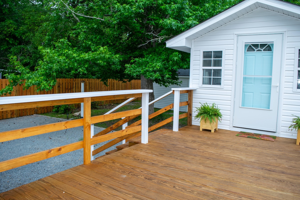 A wooden deck extends from the entryway of the cottage, featuring a white railing and potted plants at the corners. The exterior door is framed by a large window, and a gravel path is visible beyond the deck, bordered by a wooden fence and lush greenery.