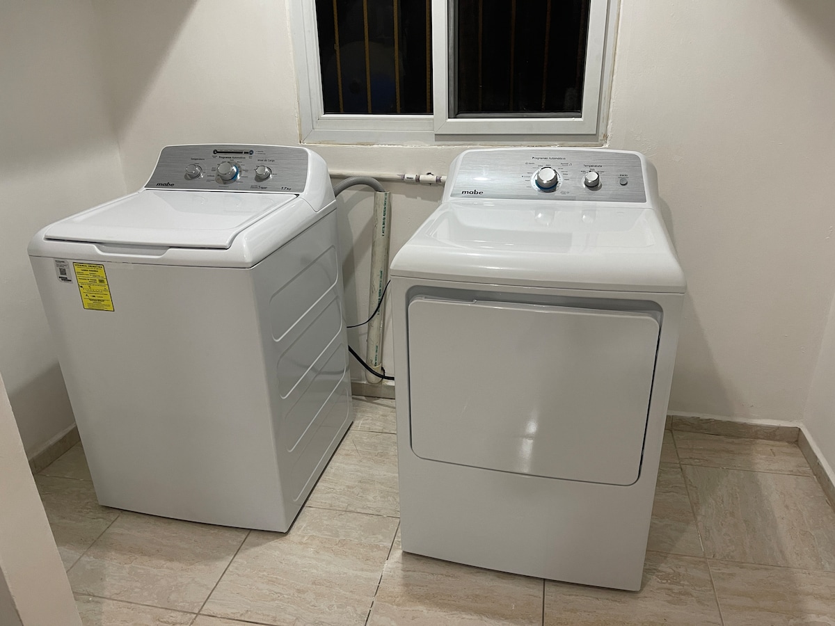 A clean laundry room features a modern washer and dryer, both in white. The appliances are positioned side by side against a neutral wall, with a window above providing natural light. Smooth tile flooring complements the space.