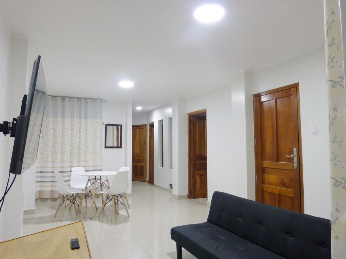 A modern and well-lit living area features a black sofa facing a wall-mounted television. A white dining table with four chairs is positioned nearby. The space is accented by light-colored curtains and hardwood doors that add warmth to the ambiance.