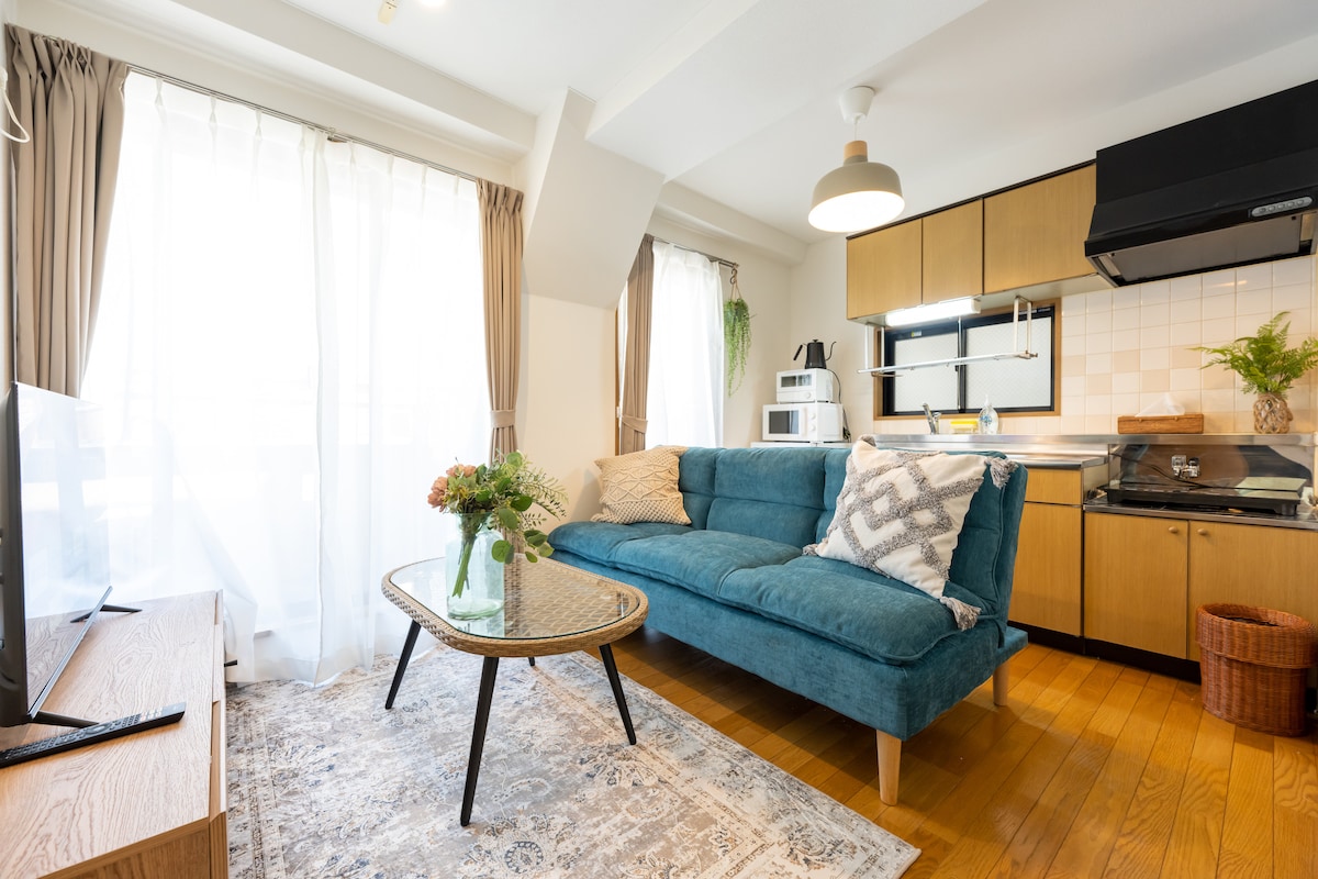A welcoming living area features a comfortable turquoise sofa adorned with patterned cushions. Natural light filters through sheer curtains, brightening the space. A wooden coffee table sits on a decorative rug, while an equipped kitchenette is visible in the background, enhancing the overall functionality.