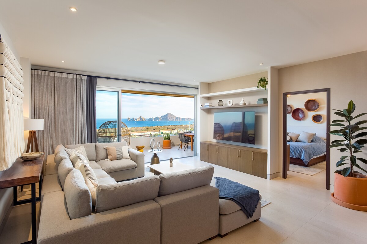 A spacious living area features a light-colored sectional sofa facing a sleek entertainment unit. Large glass doors offer views of mountains and ocean. Natural light floods the room, which includes decorative accents and a potted plant for added texture.