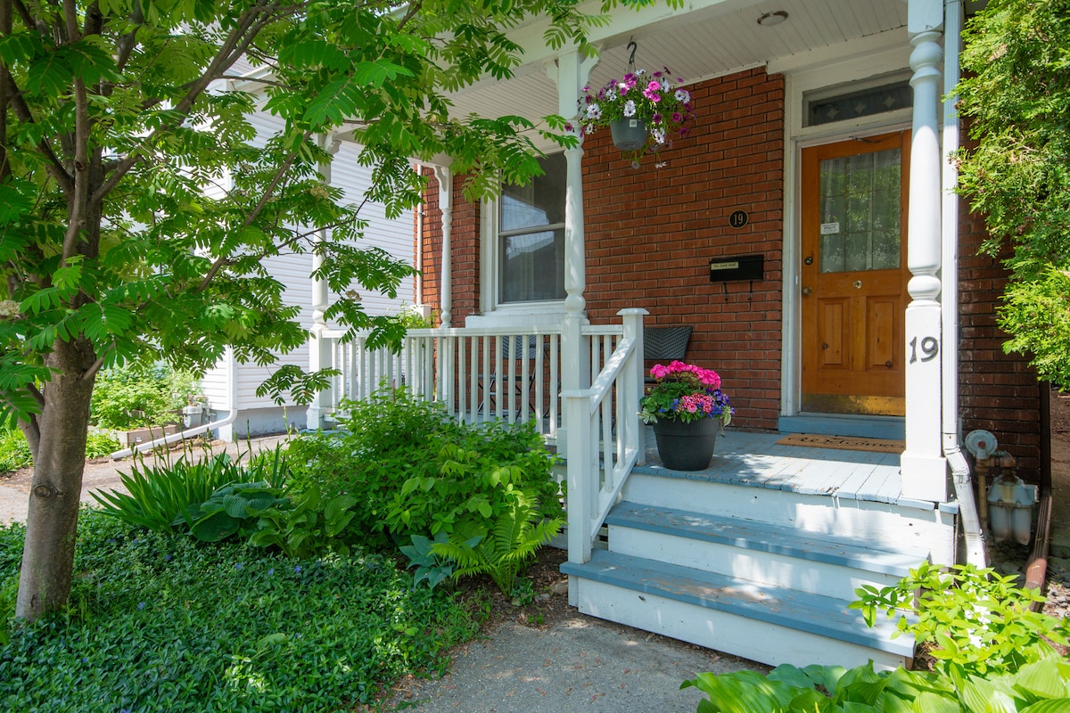 A welcoming entrance is highlighted by a wooden front door, accompanied by a mailbox. Lush greenery frames the steps, while vibrant flowers in pots add a touch of color. The porch is partially covered, offering a shaded area with a classic railing design.