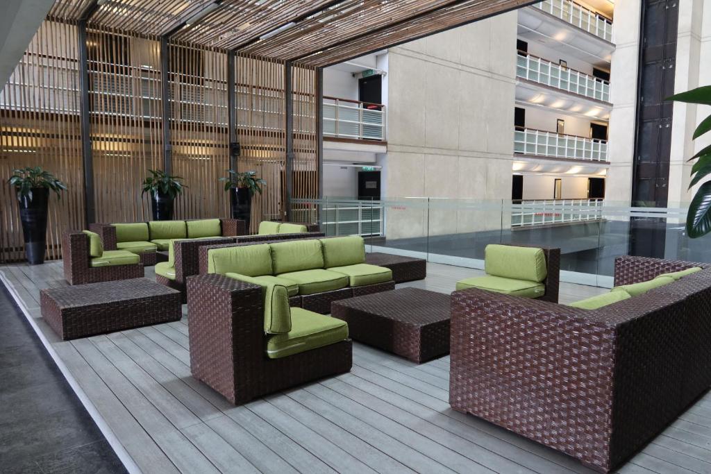 A modern lounge area features multiple seating arrangements consisting of brown wicker furniture with green cushions. A wooden deck creates a warm ambiance, while plants are positioned strategically around the space. Light pours in from the surrounding glass architecture, enhancing the openness.