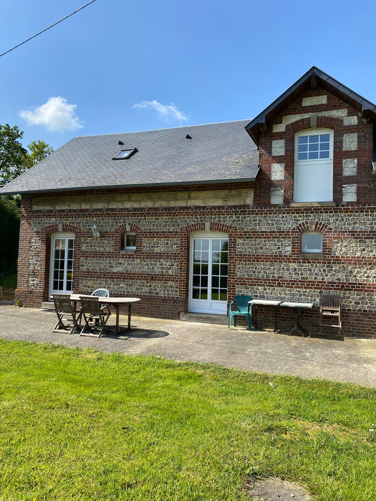 A charming brick house features a sloped roof and multiple windows, allowing natural light to fill the interior. A patio area is visible, equipped with a table and chairs, set against a backdrop of lush green grass and clear blue skies.