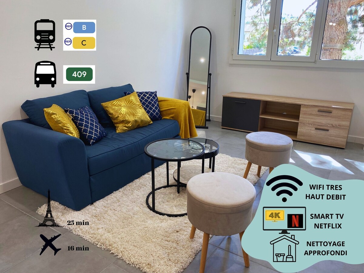 A cozy living area features a blue sofa adorned with decorative cushions. Two round tables sit on the gray tiled floor, accompanied by two soft stools. A full-length mirror reflects the natural light streaming through the large window, while a media console offers ample storage.