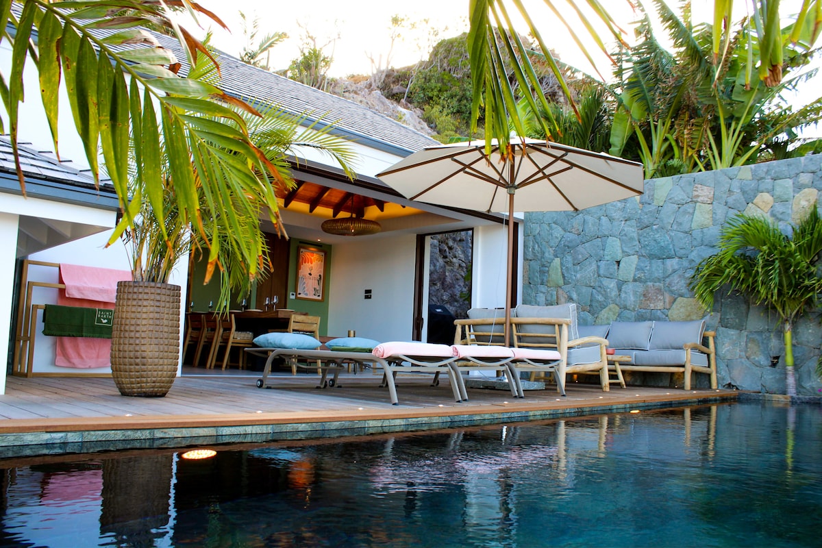 An outdoor area features a refreshing pool surrounded by palm foliage. Sun loungers are positioned neatly along the wooden deck, alongside a shaded seating area with comfortable cushions. A large umbrella provides protection from the sun, while a stone wall enhances the tropical ambiance.