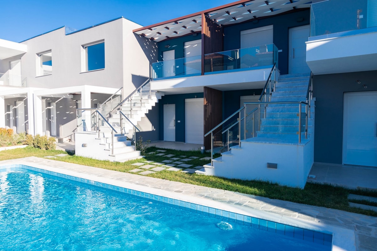 A modern two-story building is positioned next to a blue swimming pool, featuring white stairs leading to the upper level. The exterior combines light gray and royal blue colors, surrounded by landscaped gardens and clean pathways that enhance the overall appeal.