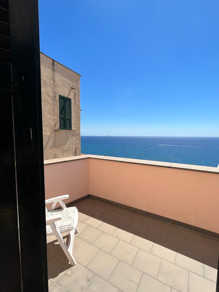 A private terrace offers an expansive view of the blue sea under clear skies. The space is complemented by a simple white chair resting on tiled flooring, inviting relaxation while enjoying the serene coastal scenery.