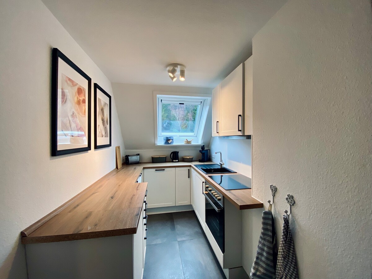 A modern kitchen is presented with light-colored cabinetry and a wooden countertop. A window allows natural light to illuminate the space, while appliances, including a stove and sink, are neatly arranged. Wall art adds a touch of decor, enhancing the inviting atmosphere.