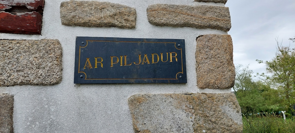 A slate plaque displaying the name 'AR PILJADUR' is attached to a textured stone wall. The wall features various sizes of natural stones, blending shades of gray and brown, with greenery visible in the background.