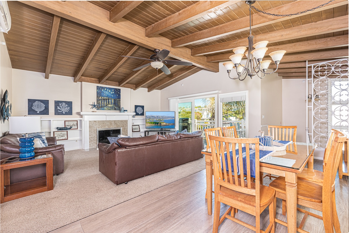 The open living space displays a vaulted wood ceiling, featuring a cozy seating area with dark sofas and a stone fireplace. A dining table with wooden chairs sits nearby, and large glass doors lead to an outdoor deck, creating a bright and airy environment.