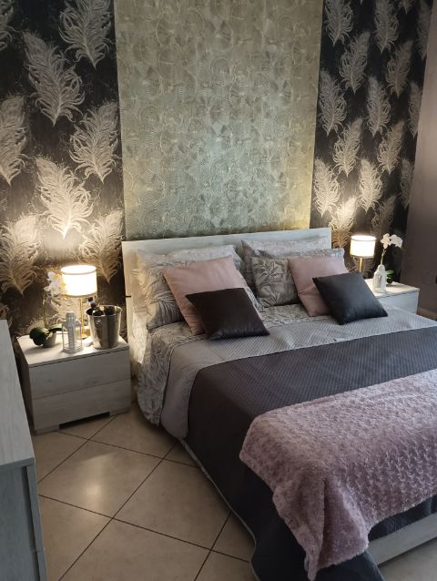 A cozy bedroom features a spacious bed with muted pillows and a soft throw. Nightstands with lamps are placed on either side, providing gentle illumination. Decorative wallpaper with a feather pattern enhances the space, complemented by a calming color palette.