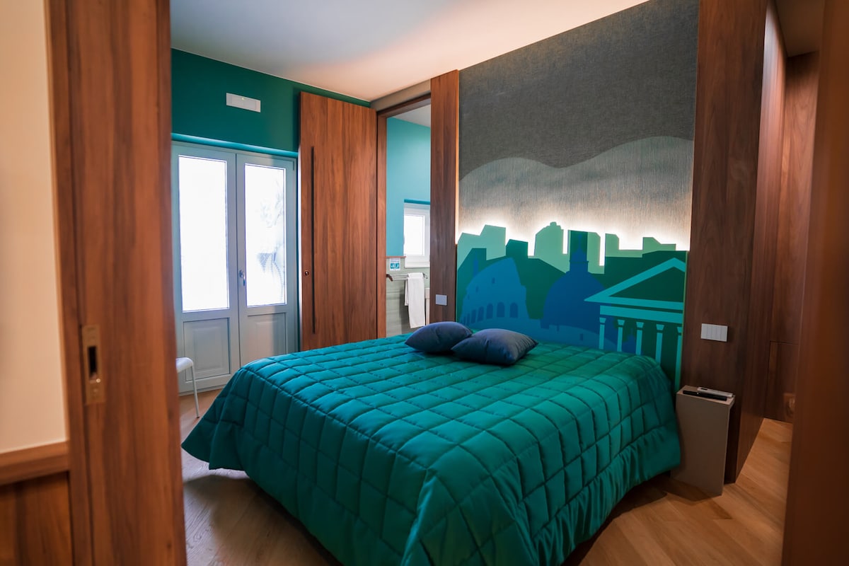 A comfortable sleeping area features a spacious bed with a textured blue-green quilt. A mural depicting a cityscape lies on the wall behind the bed, complemented by soft lighting. Natural light enters through two large windows, enhancing the cozy environment.