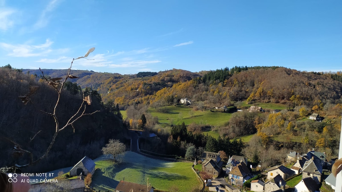 A picturesque view of lush green valleys and rolling hills under a clear blue sky is presented. Leafless trees and scattered homes with stone facades are visible, set against a backdrop of golden autumn foliage covering the hills. The peaceful landscape conveys a sense of tranquility.