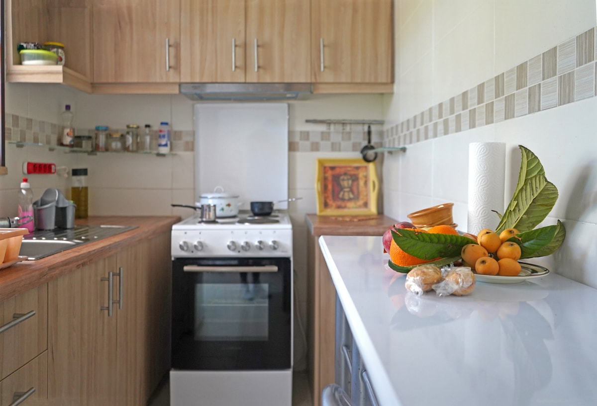 A well-organized kitchen features wooden cabinetry and light-colored countertops. A stove and oven are positioned near a sink, with various kitchen items and condiments visible on the shelves. A selection of fresh fruit and pastries is artfully arranged on the countertop.