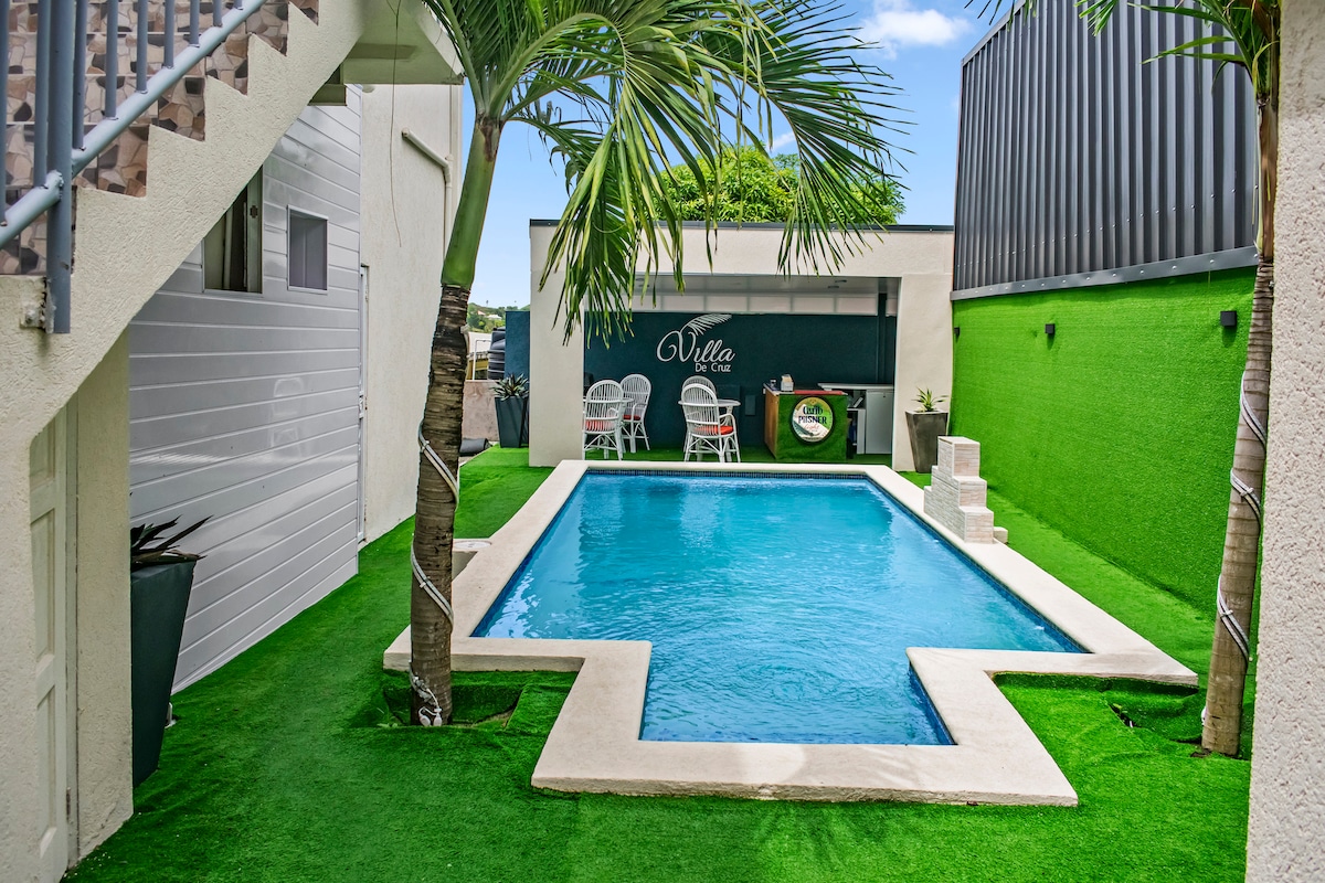 A serene shared yard features a luxurious swimming pool surrounded by vibrant green grass. Two white chairs are positioned near the pool, offering space for relaxation. The area is framed by palm trees, contributing to a calming outdoor atmosphere.