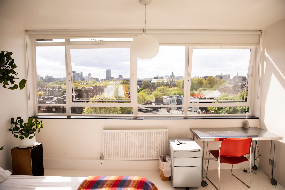 A bright room features large windows that offer expansive views of central London. A spacious bed is accompanied by a minimal desk and chair, while green plants add a touch of nature. Soft light floods the space, highlighting the clean design.