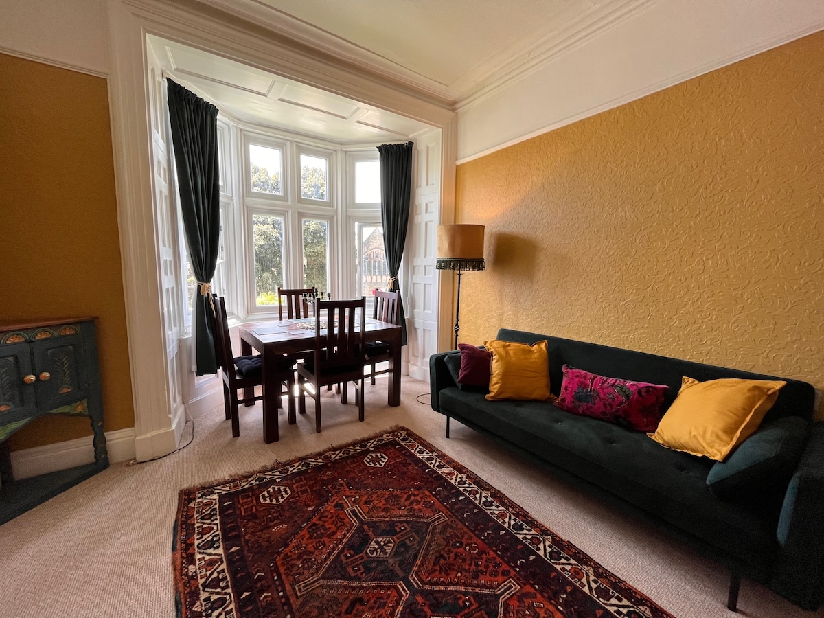 A spacious lounge area features a dark green sofa adorned with colorful cushions. A dining table with four chairs is positioned near the prominent bay windows, allowing natural light to fill the room. Soft lighting is provided by a floor lamp next to the sofa.