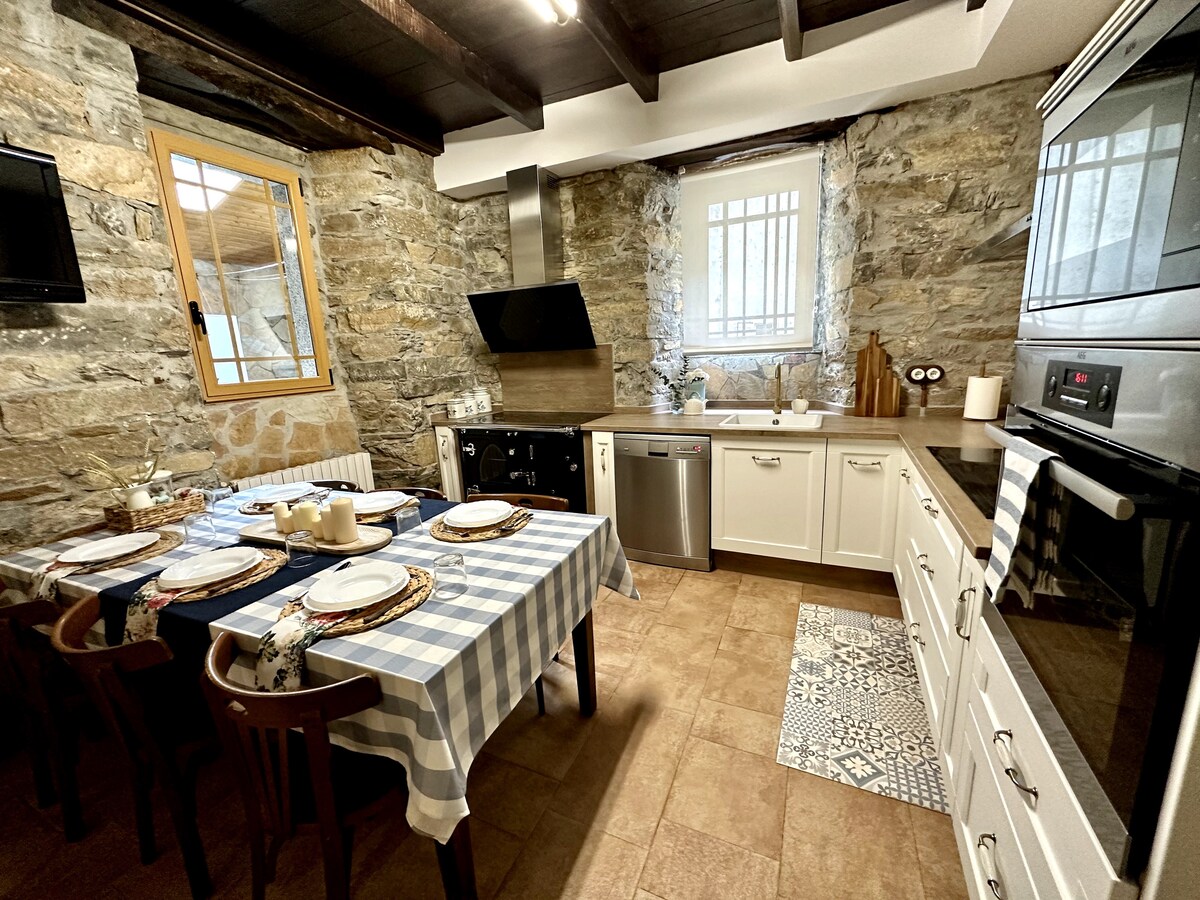 A rustic kitchen with stone walls and wooden beams features a wooden dining table set with checkered tableware. Modern appliances, including a silver dishwasher and black stovetop, are present, alongside a spacious countertop. Natural light pours in through a window, enhancing the cozy atmosphere.