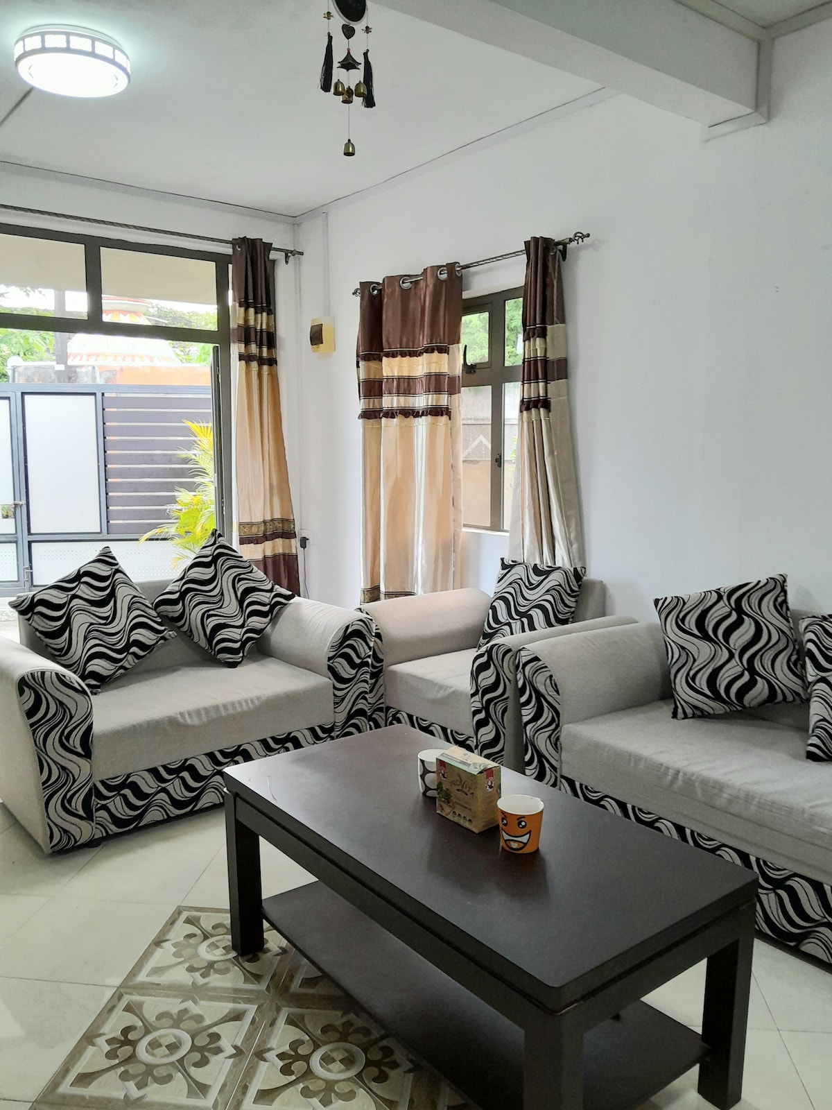 The living room features a neutral color palette with patterned sofas and a coffee table. Large windows allow natural light to enter, while the curtains provide a soft contrast. A decorative rug adds texture to the tiled floor, creating a welcoming space.