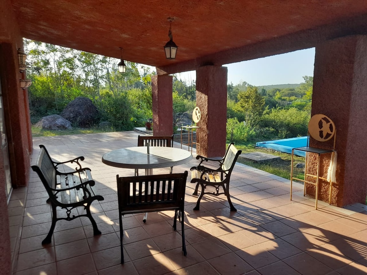 A spacious outdoor patio features a round table surrounded by wooden chairs. The area is shaded by a roof, with views of greenery and a pool beyond. Soft natural light illuminates the tiled floor, creating a serene atmosphere.