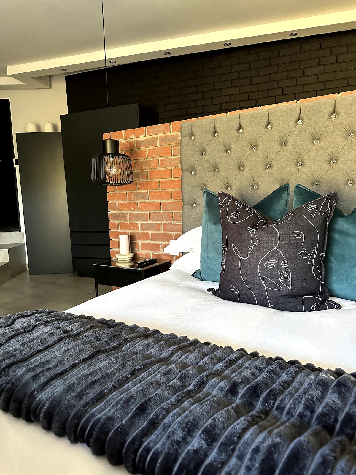 A stylish room features a plush bed adorned with decorative pillows against a textured grey headboard. A complementary throw rests across the bed, while modern lighting accents the space. The backdrop includes a striking brick wall, contributing to the room's contemporary atmosphere.