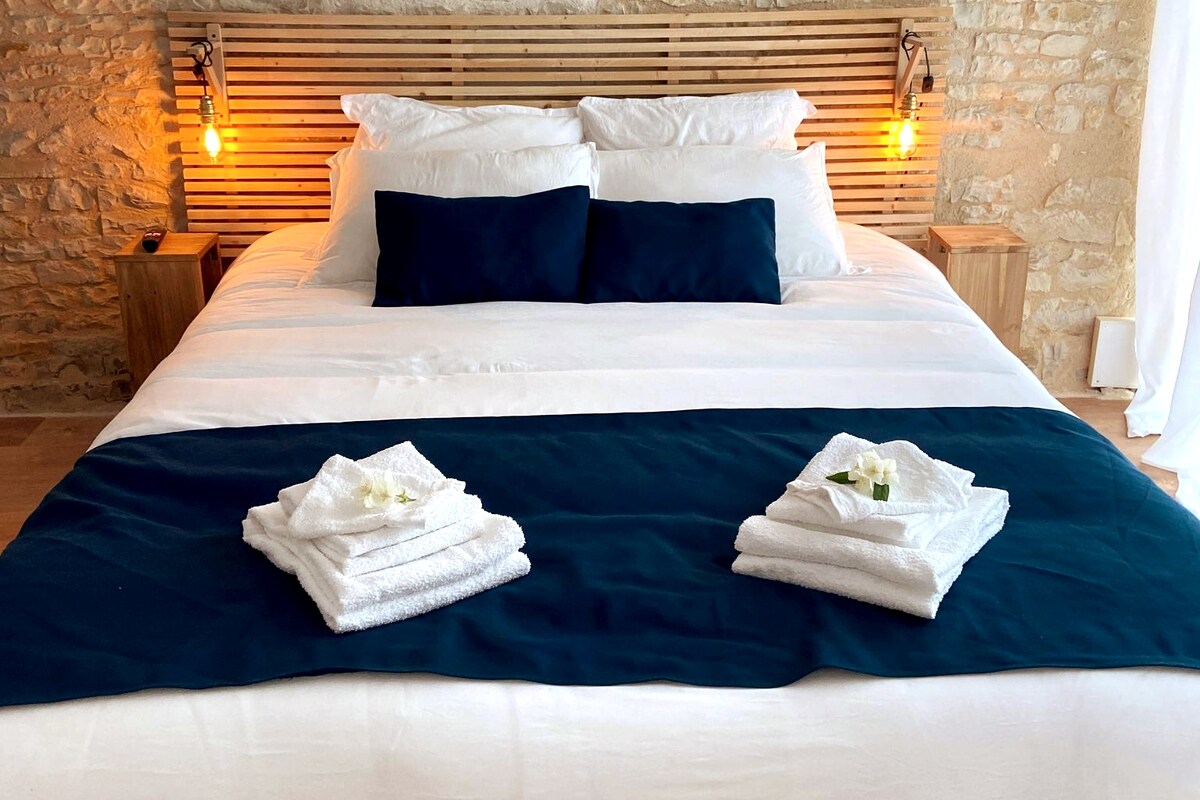 A spacious bed is centered against a rustic stone wall, featuring light-colored linens and dark blue accents. Two decorative pillows enhance the bedding. White towels are neatly arranged at the foot of the bed, accompanied by small decorative elements for a fresh touch.