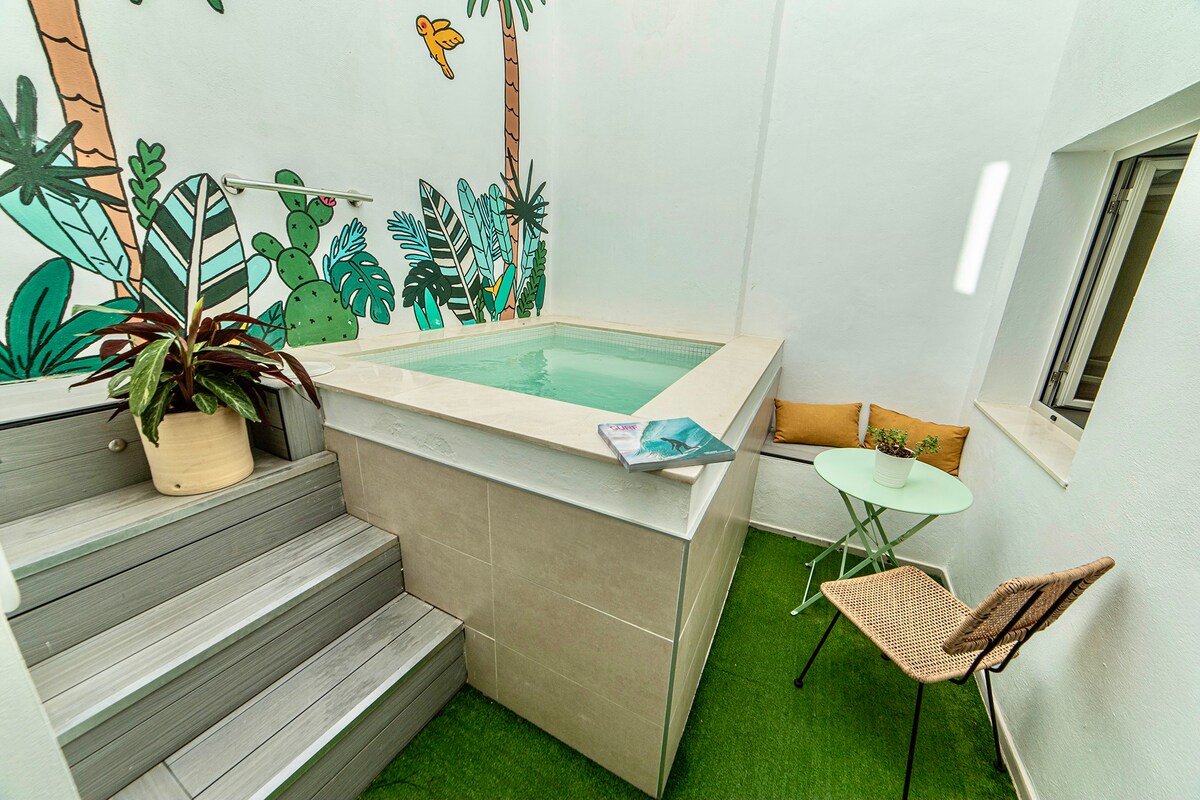 A private pool is situated outdoors, surrounded by tropical-themed wall art. Steps lead up to the pool, which features a smooth stone edge. A small green table and a chair are positioned nearby on artificial grass, offering a serene spot for relaxation.