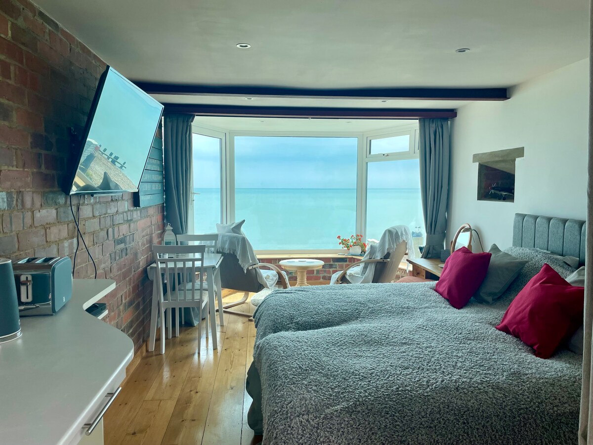 An open-plan studio apartment features two beds dressed in cozy linens, flanked by vibrant red pillows. A bay window offers panoramic sea views, complemented by a small dining table and chairs nearby. A wall-mounted TV and kitchenette are visible, enhancing the room's functionality.
