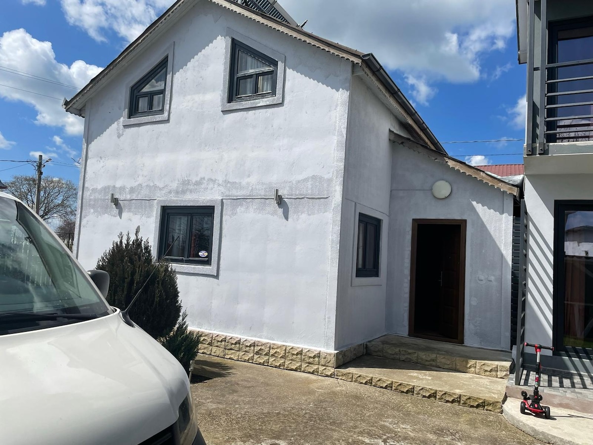 The exterior of the villa features a whitewashed facade with multiple windows, providing a bright appearance. A small entrance is visible, along with a concrete path leading up to the door. The surrounding area includes neatly trimmed shrubs and parking space.