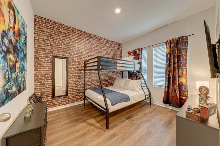 Cozy child’s or guest bedroom with a faux brick accent wall and fun fantasy-themed decor. Features a metal bunk bed with white and blue bedding, light laminate floors, patterned curtains, and a flat-screen TV for entertainment.