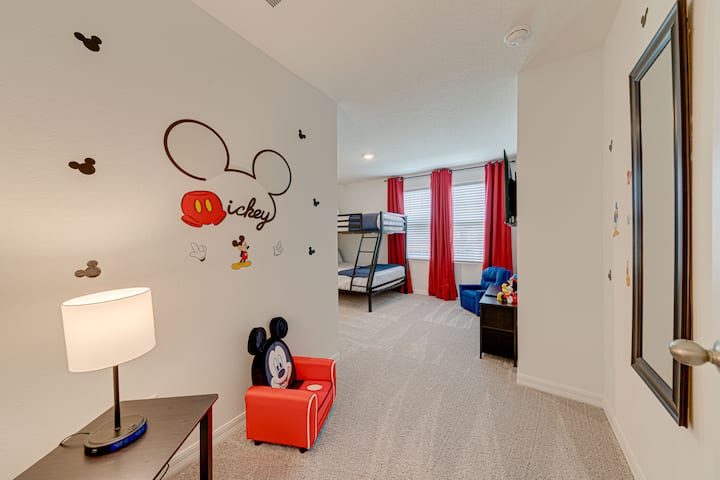 Bright child’s bedroom with fun Mickey Mouse-themed walls and decor. Features a bunk bed with red curtains, a red chair, dark blue armchair, and matching end tables. Neutral walls and carpet create a warm, inviting space for play and rest.