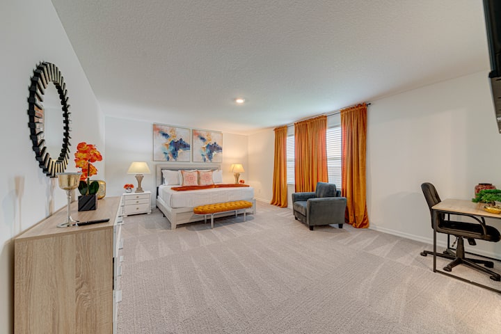 Bright master bedroom with a queen bed, mustard bench and curtains, a cozy gray armchair, and a small desk area—perfect for relaxing and working.