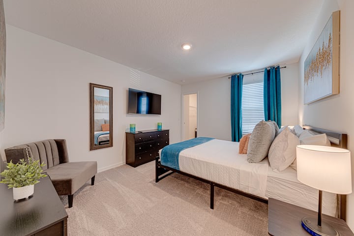 Bright, modern bedroom with teal accents, a mounted TV, and a cozy seating area. Natural light highlights the clean, inviting design—perfect for restful stays.