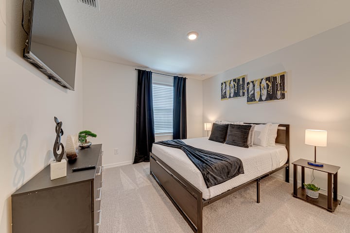 Modern master bedroom with a king bed in dark gray-brown tones, white sheets, and cozy blankets. Features dark curtains, a wall-mounted TV, a dark dresser with simple decor, and light beige-gray carpet for a clean, calm vibe.