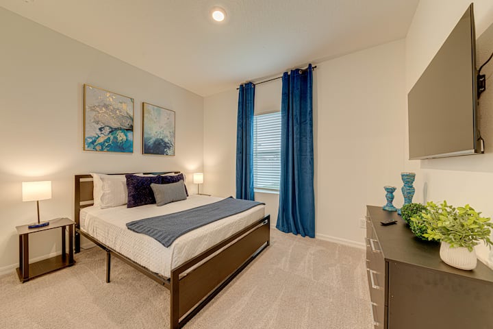 Spacious master bedroom with a dark brown metal-frame bed, white bedding, and blue accent pillows and throws. Gold-framed abstract art hangs above the bed. A wall-mounted TV sits above a window with dark-blue velvet curtains and light-brown blinds.