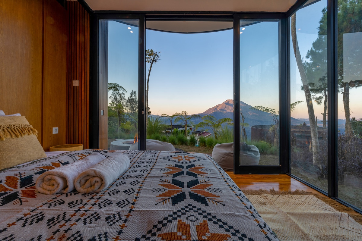 A bright bedroom features large glass walls that provide expansive views of nearby mountains. The bed is adorned with a patterned blanket, and rolled towels are neatly arranged on top. Soft lighting enhances the welcoming atmosphere in this modern minimalist space.
