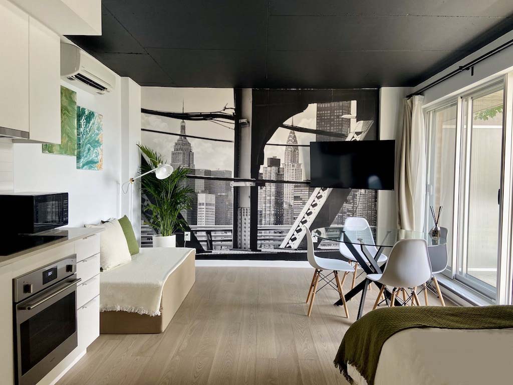 A modern studio showcases a striking black-and-white cityscape mural on one wall. A cozy seating area with a light-colored sofa is positioned near a glass dining table surrounded by contemporary chairs. Large windows provide natural light, enhancing the open feel of the space.