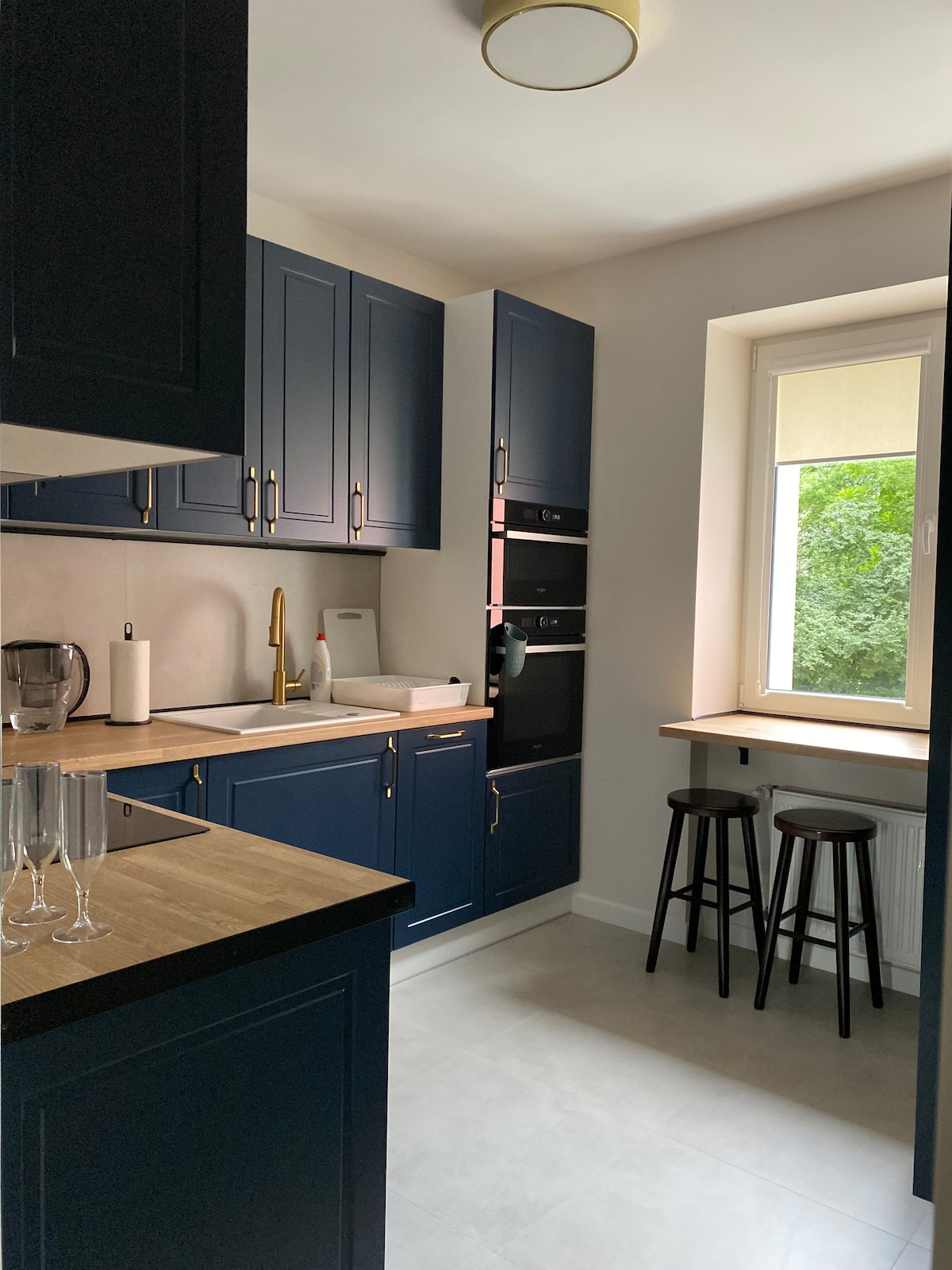 The kitchen features sleek navy cabinetry with modern gold fixtures. A small table is positioned by the window, complemented by two black stools. Natural light filters through the window, enhancing the inviting and functional space.