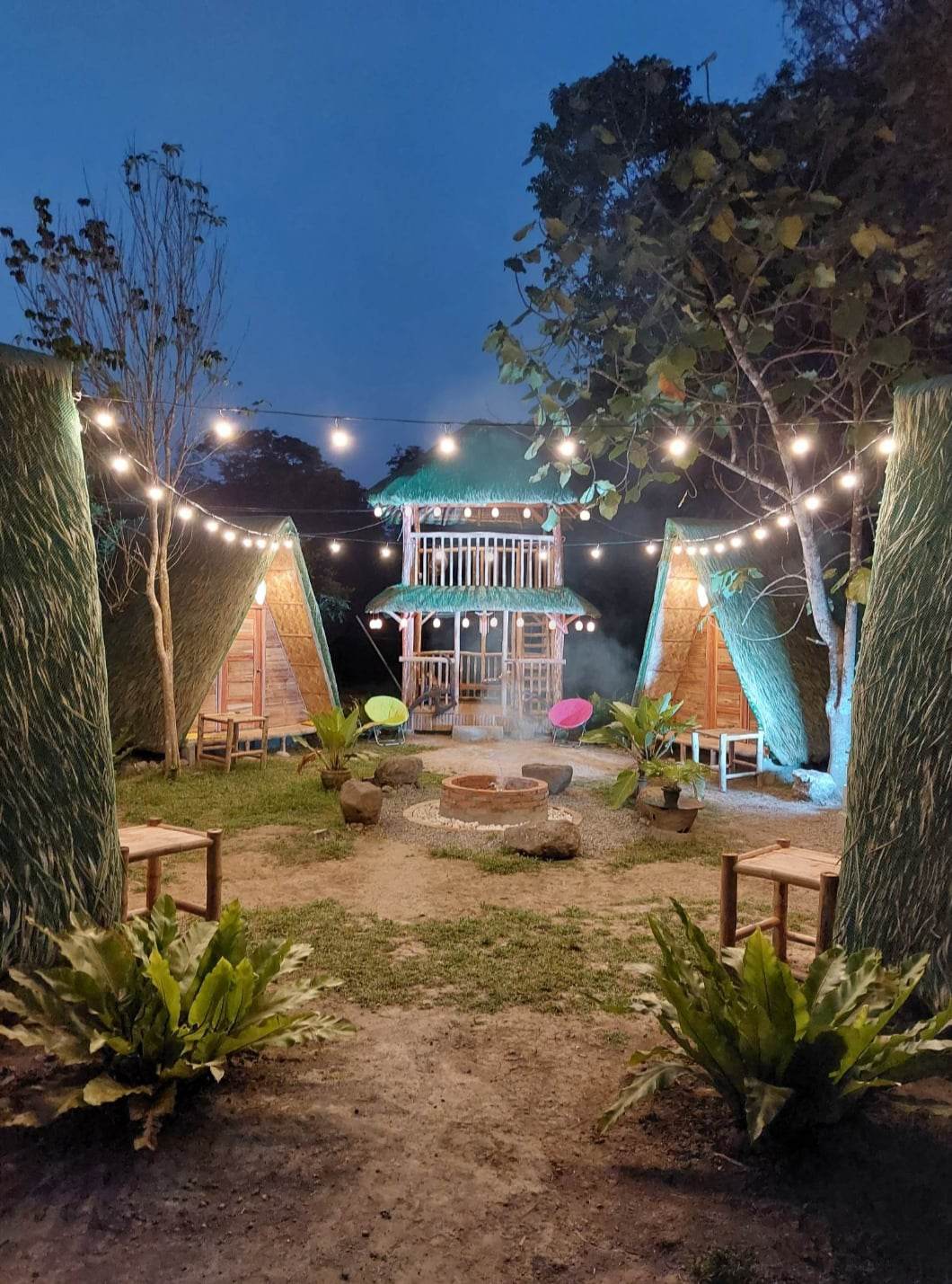Two traditional bamboo cottages are positioned on either side of a central gathering area. Soft lighting from string lights illuminates the scene, highlighting the inviting fire pit surrounded by seating. Lush greenery adds to the tranquil ambiance of this outdoor space.