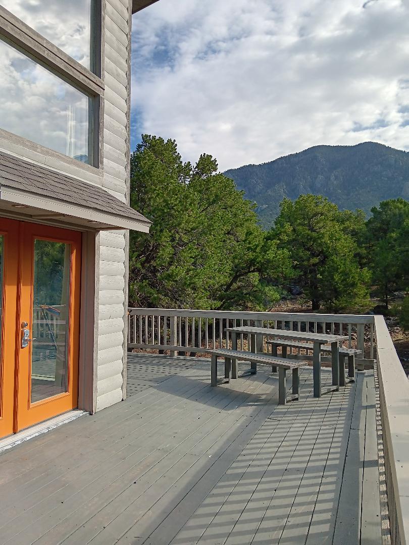 A spacious deck features a long, wooden table with attached benches, inviting outdoor relaxation. The vibrant orange doors provide access to the interior, with expansive views of surrounding greenery and distant mountains, enhancing the connection with nature.
