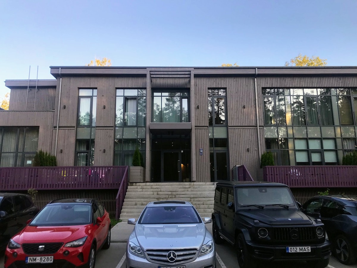 The exterior of a modern two-story apartment building is displayed, featuring large glass windows and a wooden facade. A set of stairs leads to the entrance, flanked by neatly trimmed shrubs. Several parked cars, including a silver sedan and a black SUV, are visible in the foreground.