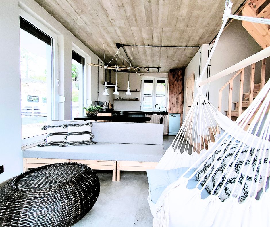 An open-concept living space showcases a cozy seating area with a low-profile sofa. A kitchen area features modern appliances and an island with pendant lighting above. A hammock hangs nearby, inviting relaxation. Natural light streams in through large windows, illuminating the concrete floor.