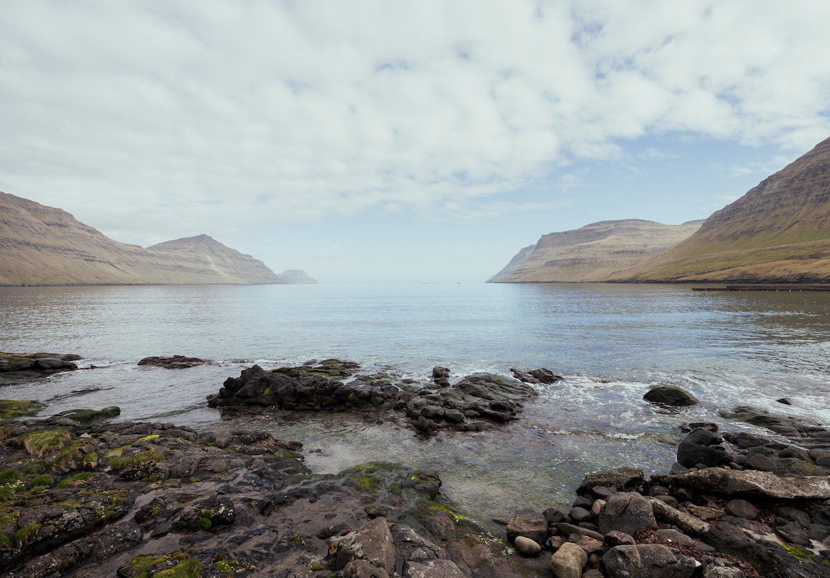 A serene coastal landscape features calm waters reflecting soft clouds above. Smooth rocks are seen along the shoreline, with grassy hills rising gently on either side. The tranquil scene invites exploration and appreciation of the natural beauty surrounding the area.
