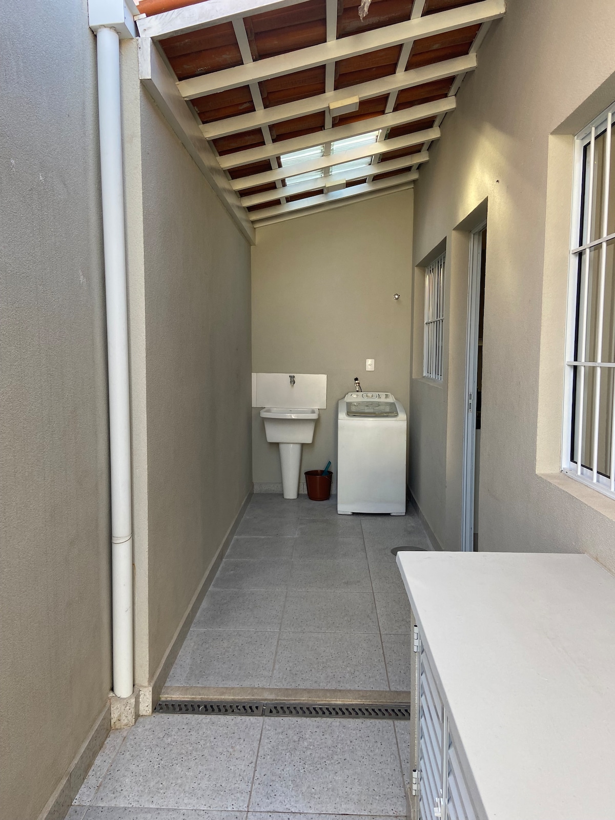 A narrow, well-lit corridor is shown, featuring a washing machine next to a sink. Natural light filters into the space through a skylight above, and light-colored tiles provide a clean and open feel. A small potted plant adds a touch of greenery.